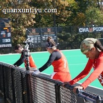 A group of athletes wearing orange uniforms are on the sideline of a sports field, resting against a fence. Behind them, a large digital scoreboard displays team names and scores. The field is bright green, and several banners with sponsor logos and championship wins are visible. The athletes appear to be taking a break, possibly before or during a field hockey game.