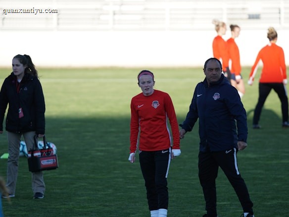 A group of people are gathered on a sports field. Some individuals are dressed in athletic gear, including a woman in a red sports jersey and a man beside her in a blue jacket, appearing to be in conversation. Additional people in similar athletic clothing are visible in the background.