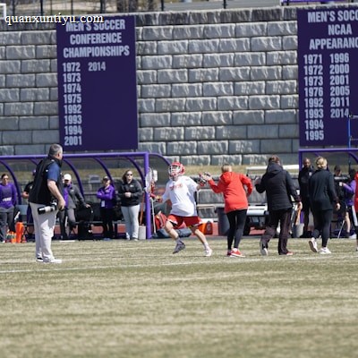 A sports team is practicing or playing on a grassy field in front of a large stone wall with banners displaying men's soccer achievements. People are wearing sports attire, and some are engaged in conversation or coaching.