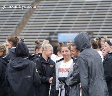 A group of people, likely athletes, are gathered together on a sports field. They are wearing black jackets with some individuals dressed in white sports uniforms underneath. A few people have their hair tied up in buns, and most are engaged in conversation. The background shows tiered stadium seats, suggesting the setting is an outdoor sports venue.