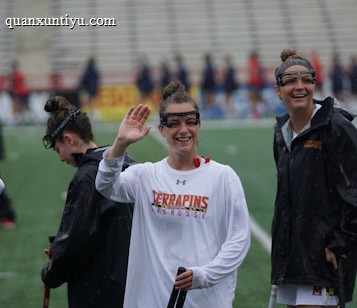 Several athletes in sports attire are on an outdoor sports field, interacting and smiling. One individual in the foreground waves with a friendly expression, while another stands beside her, also smiling. They wear protective goggles and gear associated with lacrosse. The background features indistinct figures in dark clothing against a backdrop of stadium seating.