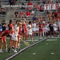 Athletes are lined up on a sports field, preparing for a game. They are dressed in athletic uniforms with some in white and others in red. The players hold lacrosse sticks and wear eye protection. In the background, spectators fill the stadium seats, mostly dressed in casual attire. A large banner is displayed behind them.