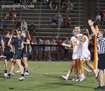 Players wearing sports uniforms are gathered on a lacrosse field. A team in dark uniforms and a team in white uniforms with one player wearing number 28 are seen celebrating near a goalpost. A referee in a striped shirt is raising an arm, indicating a call or score. Spectators are visible in the stands behind.