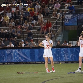 Athletes wearing white sports uniforms stand on a field with lacrosse sticks on the ground. They appear to be part of a lacrosse game, and a large audience is visible in the background seated on bleachers. The environment suggests a competitive sports event.