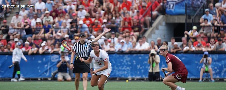 A sports event with two athletes engaged in a lacrosse game. One athlete in a white jersey is holding a lacrosse stick, appearing to prepare for a play. A referee in a striped uniform is nearby. The background features a large audience seated in a stadium, with many spectators wearing bright and casual clothing. Some photographers are also visible near the field.