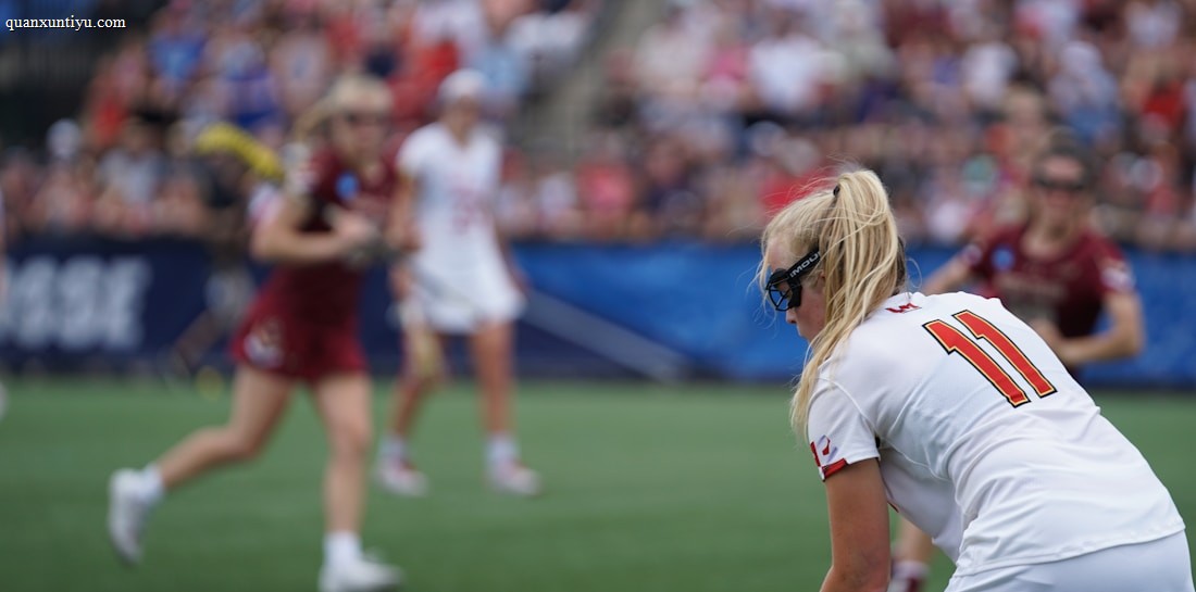 A sporting event with athletes on a grass field, one in a white jersey with the number 11 in the foreground, holding a lacrosse stick. Players in the background wear maroon uniforms. Spectators fill the stands creating a lively atmosphere.