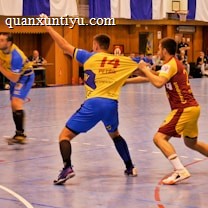 A dynamic indoor sports scene with players in motion on a court. Two athletes in blue and yellow uniforms are engaged in an intense handball game, with one player gripping the other's jersey. The background shows other players, several seated people, and advertisements on the walls.