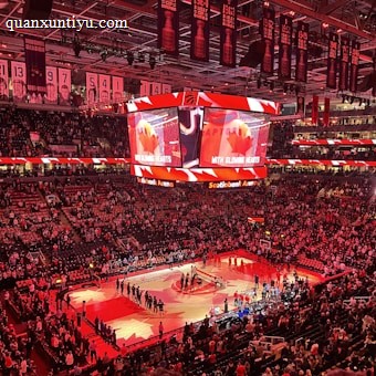 An indoor sports arena filled with spectators and a basketball court illuminated by red lighting. The scoreboards and video screens display promotional content, with numerous banners and championship flags hanging from the ceiling. Players and officials are gathered on the court, possibly in preparation for a game or event.